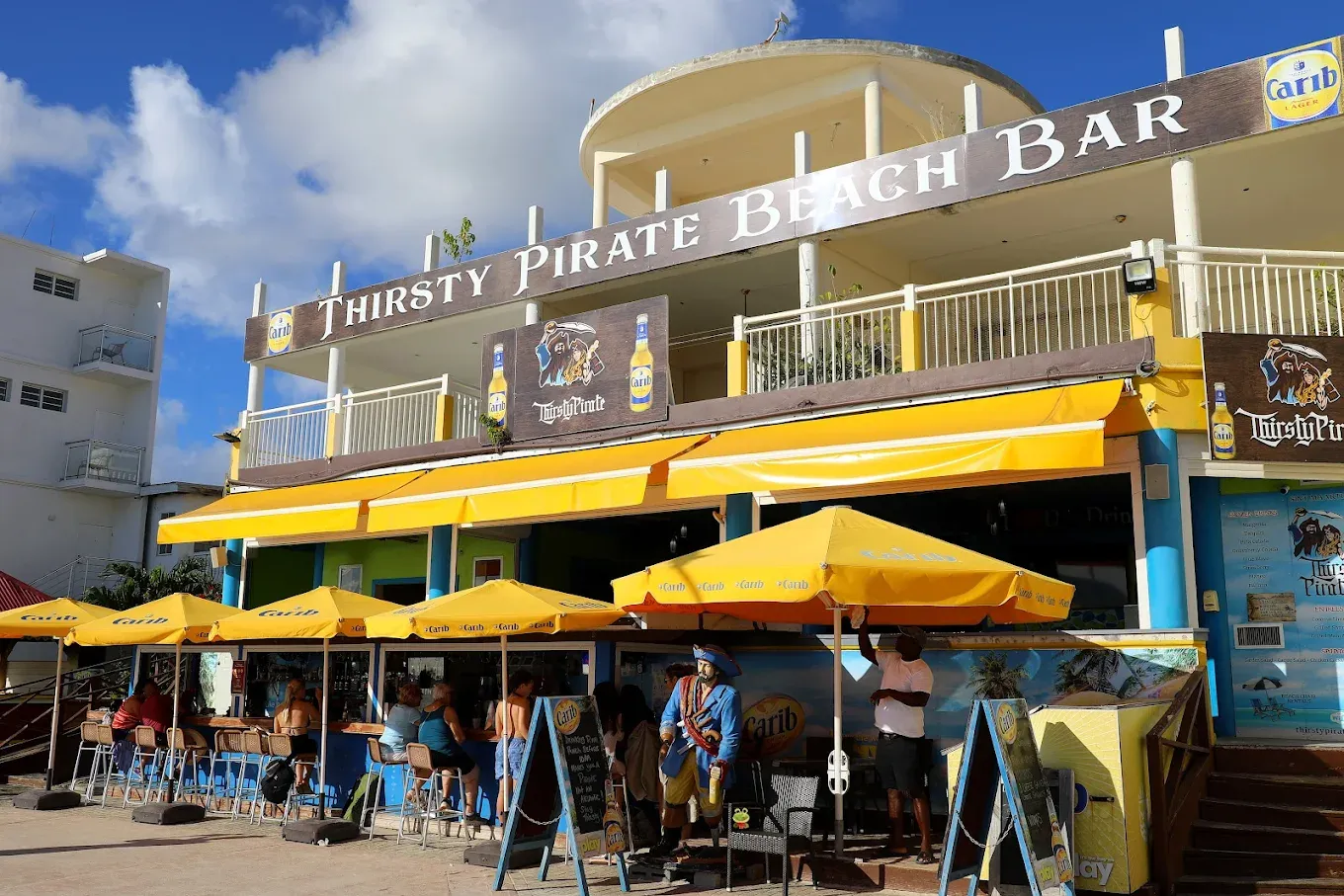 Thirsty Pirate Beach Bar storefront with its yellow awnings, pirate statue and guests at the open-air Front Street bar in Philipsburg